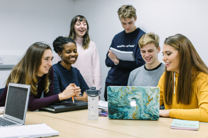 Six students are smiling looking at notebooks and a laptop screen.