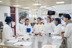 School students standing in a food science lab in lab coats with equipment and pieces of paper laid out in front of them.